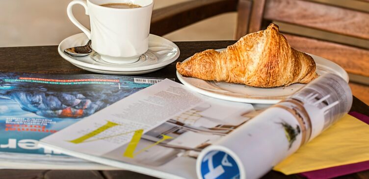 Table at a cafe with two print magazines and a cup of coffee and a croissant.