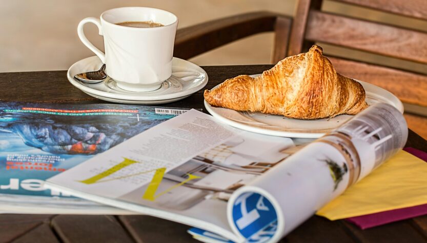 Table at a cafe with two print magazines and a cup of coffee and a croissant.